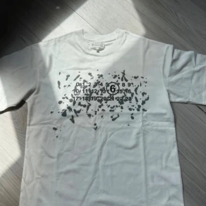 Vit t-shirt Maison Margiela med tryck - Snygg vit t-shirt från Maison Margiela med svart grafiskt tryck och siffror på bröstet. Klassisk rund hals och korta ärmar. Materialet är mjuk bomull som känns skönt mot huden. Perfekt för dig som gillar streetstyle och unika designer.