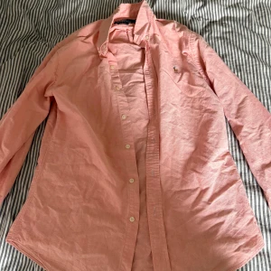 Rosa skjorta från Polo Ralph Lauren - Säljer en snygg rosa skjorta från Polo Ralph Lauren i slim fit. Skjortan har klassisk krage, långa ärmar och knäppning framtill. Ikoniska broderade loggan på bröstet. Perfekt för dig som gillar stilrena och fräscha plagg.