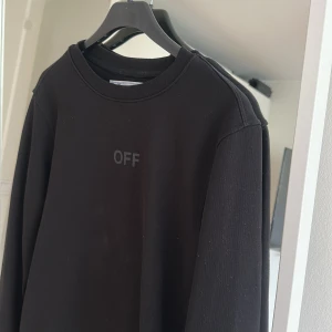 Svart sweatshirt från Off-White - Svart sweatshirt från Off-White med diskret 'OFF'-tryck framtill och stora svarta diagonala ränder på ryggen. Klassisk rund halsringning och långa ärmar. Tillverkad i mjuk bomull, perfekt för en clean streetwear-look.