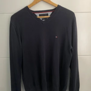 Mörkblå v-ringad tröja Tommy Hilfiger - Snygg mörkblå v-ringad tröja från Tommy Hilfiger i premium bomull. Klassisk design med liten broderad logga på bröstet och ribbade muddar vid hals, ärmslut och nederkant. Perfekt för dig som gillar stilrena och tidlösa plagg.