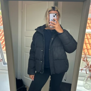Svart pufferjacka från 157, XS/S - Säljer en svart pufferjacka från 157 i storlek XS/S. Jackan har huva, dragkedja och knappar framtill samt dragsko nertill. Den är quiltad och har en loose passform, perfekt för kalla dagar. Lite sminkmärken på jackans insida.