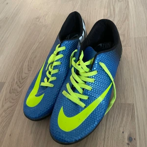 Nike Mercurial blå fotbollsskor - Nike Mercurial fotbollsskor i blått med neongula detaljer och snörning. Skorna har en texturerad syntetisk ovandel och svart häl med vit logga. Sulan har hexagonmönster och neongula partier för extra grepp på planen. Perfekt för dig som vill sticka ut på fotbollen.
