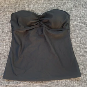 Svart bandeau  - Snygg svart bandeau tankini med lätt rynkad byst och smickrande passform. Toppen är axelbandslös och har en diskret snörning framtill samt spänne i ryggen. Perfekt för strand eller poolhäng, enkel att matcha med olika underdelar.