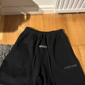 Svarta shorts Essentials Fear of God - Svarta shorts från Essentials Fear of God med elastisk midja och snörning. Mjukt bomullsmaterial, normal passform och diskret logga framtill. Perfekta för chill dagar eller streetwear-stil. Bakficka för extra funktion.