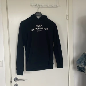 Svart hoodie från Peak Performance - Säljer en svart hoodie från Peak Performance med vit logga på bröstet. Klassisk modell med huva och magficka, perfekt för chill dagar eller när du vill ha en enkel och snygg look. Långärmad och mjuk insida.