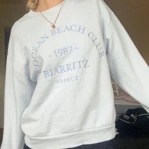 Ljusblå sweatshirt från Gina Tricot - Supermjuk ljusblå sweatshirt från Gina Tricot med tryck 'Ocean Beach Club 1987 Biarritz France' i blått framtill. Tröjan har rund hals, ribbade muddar och en relaxed fit som är perfekt för chill dagar.