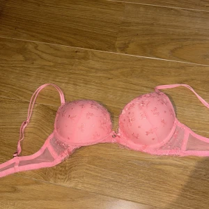 Rosa blommig spets-bh med rosett - Supersöt rosa bh med blommigt broderi och spetsdetaljer. Kuporna är vadderade och har en liten rosett med pärla i mitten. Axelbanden är justerbara och tyget är mjukt och bekvämt. Perfekt för dig som gillar romantiska och gulliga underkläder.