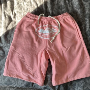 Rosa shorts från Corteiz - Snygga rosa shorts från Corteiz i mjukt material med vit snörning i midjan och tryckt logga framtill. De har elastisk midja, sidofickor och en bakficka med knapp. Perfekta för chill dagar och har en loose passform. Köpta från en kompis så osäker på om det är äkta, köp på egen risk helt enkelt! Helt oanvända iallafall då jag inte längre gillar de.
