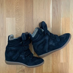 Svarta wedge sneakers med kardborre - Snygga svarta sneakers med högt skaft och inbyggd kilklack. Skorna har flera breda kardborreband och är gjorda i mocka med detaljer i skinn. Yttersulan är i gummi med brun ton. Perfekta för dig som vill ha streetstyle med extra höjd.