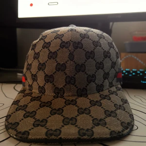 Gucci keps med GG-mönster och röd-blå rand - Säljer en snygg keps från Gucci med klassiskt GG-mönster i beige och svart. Kepsen har en röd och blå rand på sidan och är tillverkad i bomullsmix. Perfekt accessoar för att lyfta din stil. Pris kan sänkas vid snabb affär öppen för byten men möts upp då