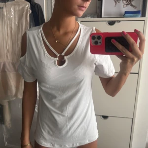 Vit topp med cut out och ringdetalj - Säljer en vit topp med coola cut out-detaljer på axlarna och en snygg ring framtill vid halsen. Toppen har korta ärmar och dubbla band över bröstet som ger en unik look. Perfekt för dig som vill sticka ut lite extra.