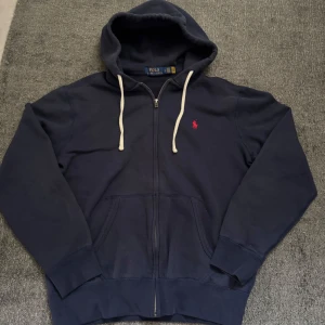 Polo Ralph Lauren Marinblå hoodie - Jag har haft hoodie i ett par månader och använt den ett få tal gånger. Hoodie är i mycket bra skick. Snygg marinblå hoodie från Polo Ralph Lauren med dragkedja och vit snörning i huvan. Tröjan har det klassiska röda Polo-logot broderat på bröstet, två fickor fram och är tillverkad i mjuk bomull. Perfekt för chill dagar och streetwear vibes. 