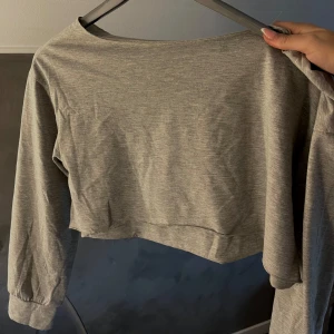 Grå croppad offshoulder topp - Säljer en ljusgrå croppad topp med offshoulder-design och långa ärmar. Toppen är i mjuk bomullsmix och har en loose passform som ger en chill vibe. Perfekt att styla med höga jeans eller kjol för en trendig look.