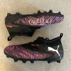 Puma fotbollsskor future 8 junior svart/lila  - Snygga fotbollsskor från Puma i svart med lila och orange detaljer. Skorna har en modern design med mönstrad ovandel, snörning och vit Puma-logga på sidan. Sulan har fasta dobbar för bra grepp på planen och ovandelen är i syntetmaterial.