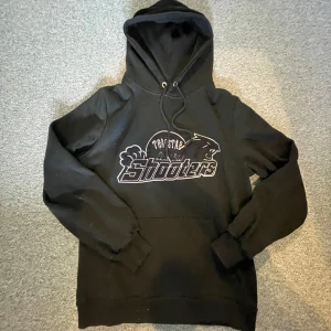 Svart hoodie Trapstar Shooters S - Svart hoodie från Trapstar med trycket 'Shooters' och pantermotiv på bröstet. Klassisk modell med huva och snörning, samt magficka framtill. Perfekt för dig som gillar streetwear och vill ha en stilren, bekväm tröja.