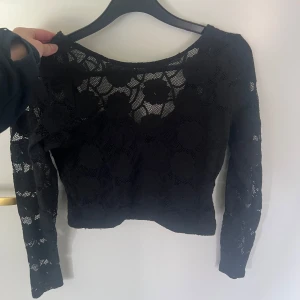 Svart spetsblus med blommönster - Croppad långärmad spetstopp. Djup rygg och svart täckande tyg över mage, rygg och bröstkorg, genomskinlig spets högst upp. Storlek xs från bikbok 