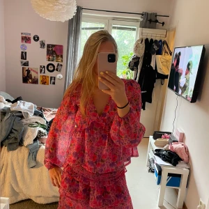 Blommigt rosa sett med kjol och blus  - Ett sett med volang kjol och blus, storlek XS på kjol storlek S på blus, från Bikbok i ett blommigt mönster med rosa, lila och röda toner. Klänningen har volangdetaljer, v-ringning och långa ärmar i ett lätt och luftigt material. Perfekt för sommarens alla tillfällen! Kan köpa enskilt 