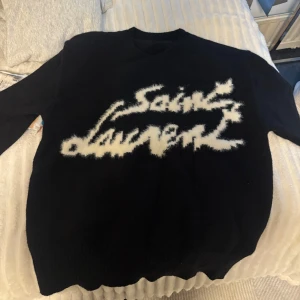 Svart Saint Laurent stickad tröja - Svart stickad tröja från Saint Laurent med vit, fuzzy logga framtill. Tröjan har rund halsringning och långa ärmar. SKIT SNYGG till hösten! Funkar både till tjejer o killar men är såld som en herrtröja.