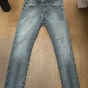 Slim Glenn ljusgrå jeans 32/32 - Ljusgrå slim fit jeans från Slim Glenn i storlek 32/32. Jeansen har en snygg tvättad look med slitning/hål på ena benet och klassiska femficksdetaljer. Perfekta för dig som gillar en smalare passform och stilren vibe.