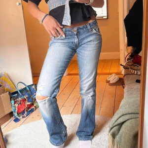 lågmidjade bootcut jeans - Snygga blå bootcut jeans från Wrangler med slitningar och hål på knät. Klassisk femficksmodell med låg midja och ljus tvätt. Jeansen har en tydlig logga på bakfickan och är tillverkade i robust denim. Perfekta för en avslappnad och cool look.