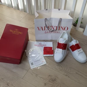 Valentino Garavani Open sneakers  - Snygga Valentino Garavani Open sneakers i vitt skinn med bred röd läderdetalj över vristen. Klassisk rund tå, platt sula och diskreta nitar baktill. Kommer med originalkartong, dustbag och påse. Perfekta för dig som vill sticka ut med stil. Säljers då dom bara ligger och samlar damm.
