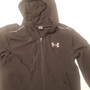 Svart jacka från Under Armour - Svart jacka från Under Armour med dragkedja och huva. Jackan har långa ärmar, snörning i huvan och logga på bröstet. Materialet är lätt och andas, vilket gör den skön att ha på sig både inne och ute. Fast pris. Inga byten. 