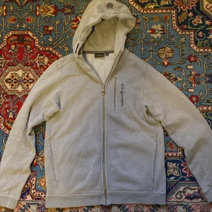 Grå hoodie från Sail Racing med dragkedja - Snygg grå hoodie från Sail Racing med hel dragkedja framtill och broderad logga på huvan. Tröjan har långa ärmar, fickor på sidorna och en liten bröstficka med dragkedja. Perfekt för chill dagar eller när du vill ha en clean streetstil.