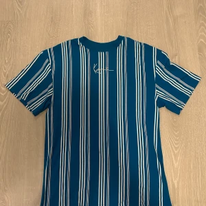 Karl Kani T-Shirt - Snygg t-shirt från Karl Kani i blått med vita vertikala ränder. Helt oanvänd utan prislapp. Väldigt neutral i passsform, lite baggy. Sitter bra över armar och midja. Om du vill diskutera pris eller något annat tveka inte på att skriva 🙏