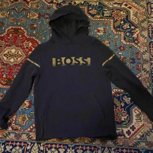 Mörkblå hoodie från BOSS med guldtryck - Snygg mörkblå hoodie från BOSS med guldiga detaljer och stort BOSS-tryck på bröstet. Tröjan har huva, långa ärmar och en klassisk känguruficka framtill. Perfekt för dig som vill ha en stilren och trendig look.