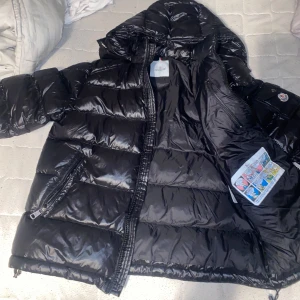 Svart Moncler pufferjacka med huva - Säljer en svart pufferjacka från Moncler med glansig finish och klassisk logga på ärmen. Jackan har huva, dragkedja framtill och två fickor med dragkedja. Insidan har en cool serietidningsdetalj. Perfekt för kalla dagar när du vill hålla stilen.