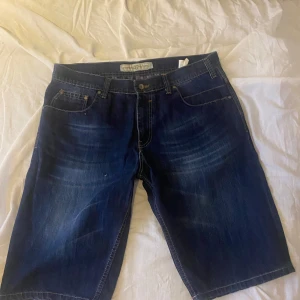 Blå jeansshorts från Fashion Factory - Snygga mörkblå jeansshorts från Fashion Factory med klassisk femficksdesign och slitningar framtill. Modellen har normal passform och är tillverkad i 100% bomull, vilket gör dem sköna och slitstarka. Perfekta för varma dagar och chill häng.