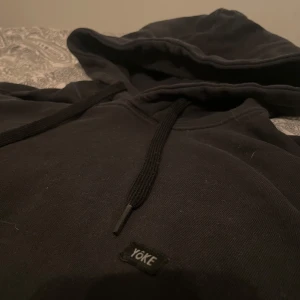 Svart hoodie från Yoke med huva - Svart hoodie från Yoke med klassisk huva och snörning. Tröjan har en stor magficka och ribbade muddar vid ärmslut och nederkant.