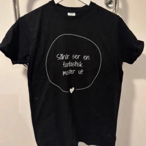 Svart t-shirt med tryck B&C Collection - Svart t-shirt från B&C Collection i storlek S med rund hals och korta ärmar. Framsidan har ett vitt tryck med texten 'Såhär ser en fantastisk moster ut' inuti en handritad cirkel och ett litet hjärta. Tillverkad i mjuk bomull, perfekt för att visa lite extra attityd.