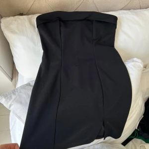 Svart axelbandslös kortklänning - Snygg svart axelbandslös klänning med figurnära passform och liten slits framtill. Perfekt för dig som gillar minimalistisk stil och vill ha en enkel men elegant look. Klänningen är kort och har en stilren design utan extra detaljer. 