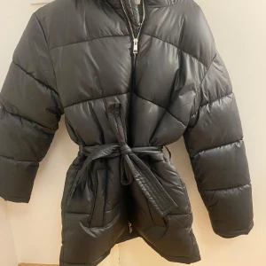 Svart pufferjacka med bälte och huva - Säljer en svart pufferjacka med huva och bälte i midjan. Jackan har quiltad design, dragkedja framtill och långa ärmar. Perfekt för kalla dagar och har en snygg oversized look. Materialet är syntet och jackan är mörk i tonen.