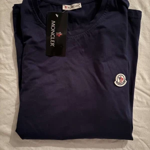 Mörkblå t-shirt från Moncler - Snygg mörkblå t-shirt från Moncler med klassisk rund hals och korta ärmar. T-shirten har Moncler-logga broderad på bröstet och är gjord i mjuk bomull som känns skön mot huden. Perfekt för dig som gillar stilrena och exklusiva plagg.                                                                                                                                                                              Nypris: 2000kr GORILLAPRIZE:1120kr            Skick:10/10 oöppnad         KVITTO MEDKOMMER    