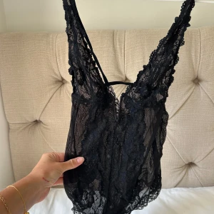 Svart spetsig body med djup v-ringning - Säljer en svart body med djup v-ringning och tunna axelband. Bodyn är gjord i transparent spets med fina broderade detaljer och har en feminin, sexig look. 