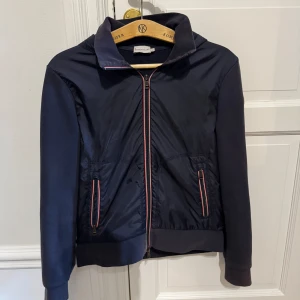 Mörkblå cardigan jacka från Moncler - Snygg mörkblå cardigan jacka från Moncler med sportig känsla. Jackan har hel dragkedja framtill, två fickor med röd-vita detaljer och ribbade muddar. Materialmix med glansig framsida i nylon och stickade ärmar. Perfekt för dig som gillar stilren och exklusiv look. Möts i Stockholm. MVH