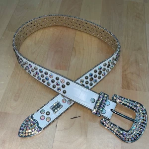BB SIMON RHINESTONE BÄLTE - Snyggt vitt bälte med massor av glittrande rhinestone-stenar och silvriga nitar längs hela bandet. Spännet är dekorerat med stora stenar som verkligen poppar. Perfekt accessoar för att sticka ut och ge outfiten extra edge.