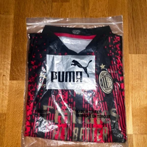 AC Milan x Puma digitalmönstrad tröja - AC Milan fotbollströja från Puma med digitalt röd-svart mönster, guldiga detaljer och klubbmärke på bröstet. Tröjan har korta ärmar, V-ringad krage och är tillverkad i lätt, ventilerande material. Perfekt för dig som vill sticka ut på planen eller på läktaren. Enda negativa är en litet tecken på slitage i kragen.
