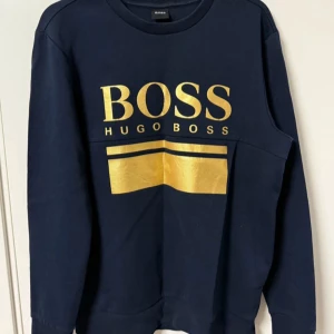 Mörkblå sweatshirt från Hugo Boss - Snygg mörkblå sweatshirt från Hugo Boss med stort guldigt tryck framtill. Tröjan har rund halsringning och långa ärmar, perfekt för en stilren och trendig look. Skön bomullskvalitet som passar till många outfits. (Använd några gånger!)