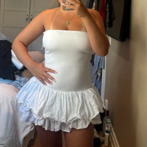 Vit volangklänning från Zara - Supersöt vit kort klänning från Zara med axelbandslös design och flera lager volanger i kjolen. Överdelen är ribbad och tight, medan kjolen har en fluffig och lekfull look. Perfekt för dig som gillar att sticka ut med en feminin och trendig stil. Den är näst intill oanvänd då jag aldrig haft tillfälle använda den