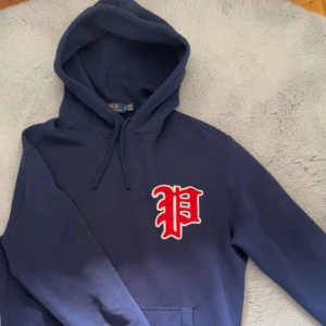Mörkblå hoodie från Polo Ralph Lauren - Snygg mörkblå hoodie från Polo Ralph Lauren med stor röd och vit P-logga på bröstet. Tröjan har huva med snörning, magficka och långa ärmar. Perfekt för dig som gillar streetwear och vill ha en klassisk college-look.