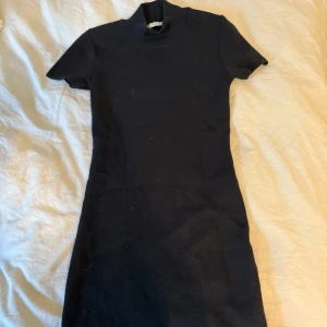 Svart stickad kortklänning - En svart stickad kortklänning med hög hals och korta ärmar. Klänningen har en enkel, rak passform och är perfekt för dig som gillar en stilren look. Materialet är mjukt och bekvämt, vilket gör den lätt att bära hela dagen.