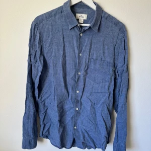 Blå linneskjorta från H&M Edition - Snygg blå skjorta i 100% linne från H&M Edition. Klassisk krage, bröstficka och knäppning framtill. Långärmad modell med avslappnad känsla, perfekt för dig som gillar stilren och enkel look. A395