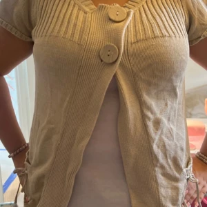 Beige stickad kofta med stora knappar - Beige stickad kofta med korta ärmar och två stora träknappar framtill. Koftan har ribbade detaljer upptill och knytband i sidorna för extra stil. Perfekt att slänga över en topp för en chill look.