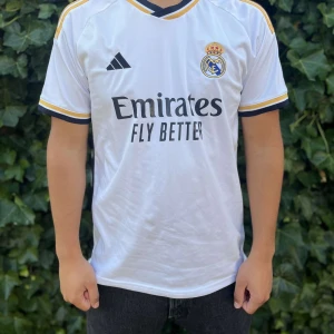 Real Madrid vit fotbollströja Adidas - Snygg vit Real Madrid fotbollströja från Adidas med klubbmärke och sponsortryck på bröstet. Tröjan har svarta och gula detaljer vid krage och ärmslut samt klassisk V-ringning. Tillverkad i lätt och ventilerande material, perfekt för match eller träning.																																																																																																																	 	Nypris:700kr GORILLAPRICE:400kr																																											SKICK:8.75/10
