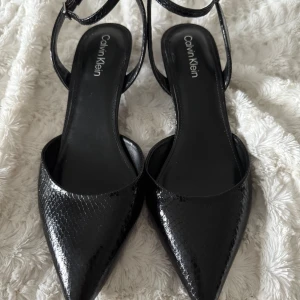 Svarta pumps från Calvin Klein - Snygga svarta pumps från Calvin Klein med spetsig tå och låg klack. Skorna har ett glansigt, ormskinnsinspirerat mönster och smala remmar runt ankeln. Perfekta för dig som vill ha en stilren och trendig look. Köpta i New York 