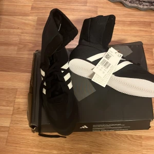 Svarta adidas boxningsskor med vita ränder - Snygga svarta boxningsskor från adidas med klassiska vita ränder på sidorna. Skorna har hög modell, snörning och en vit, greppvänlig sula. Ovandelen är i mesh och syntet för bra ventilation och lätt känsla. Perfekta för dig som vill ha stilrena och funktionella träningsskor. Oanvända skor, helt nya.