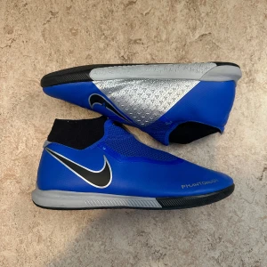 Nike Phantom Vision blå fotbollsskor - Nike Phantom Vision fotbollsskor i blått med silverdetaljer och svart sula. Skorna har en strumpliknande passform, meshpaneler och stora svarta swoosh-loggor. Ovandelen är i syntetmaterial och designen är modern och sportig, perfekt för inomhusspel.
